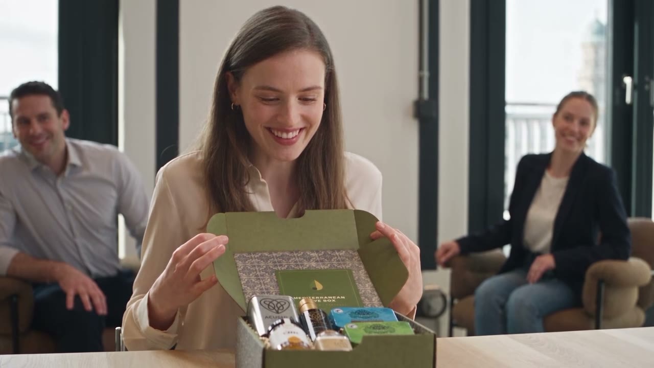 Lächelnde Mitarbeiterin öffnet im Besprechungsraum die Olive Box, zwei Kollegen schauen zu